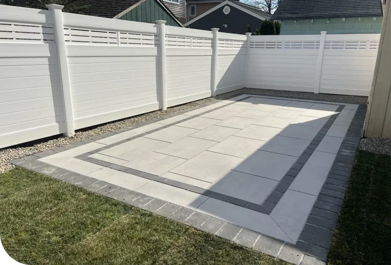 A backyard area in Jackson Township with white privacy fencing features a rectangular hardscaping patio of large light stone tiles bordered by gray bricks, surrounded by gravel and a patch of grass in the foreground.