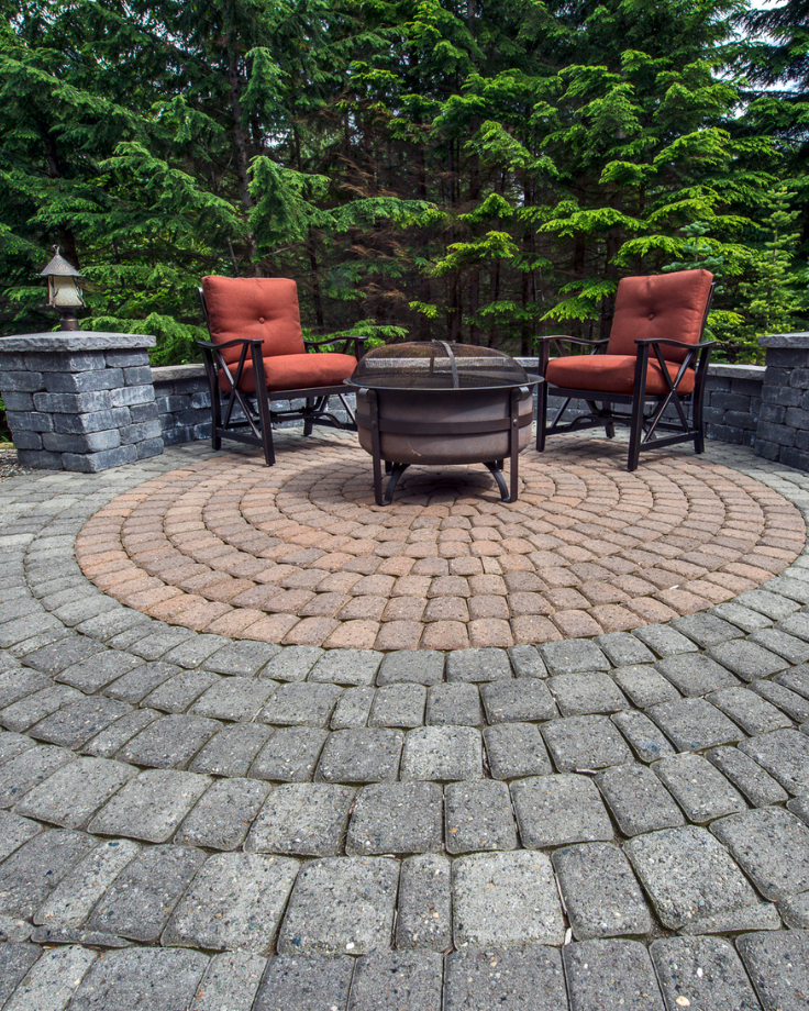 A circular stone patio with two cushioned chairs and a fire pit, surrounded by a low stone wall and green trees—expertly crafted with Landscaping Services in Shrewsbury.