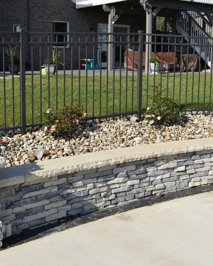 A stone retaining wall topped with gravel and small rose bushes sits in front of a metal fence, showcasing expert Landscaping Services in Shrewsbury, with a green lawn and patio area visible in the background.