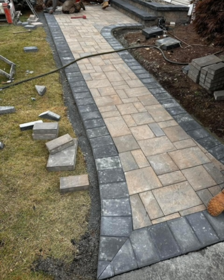 A stone walkway under construction with light and dark rectangular pavers; some stones and construction tools are scattered on the adjacent grass and soil, part of ongoing Landscaping Services in Shrewsbury.